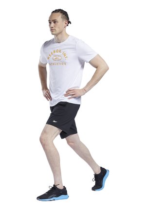 Camiseta deportiva blanca con texto naranja, pantalones cortos negros y zapatillas negras con acentos azules. El modelo está en una posición de caminar posada.
