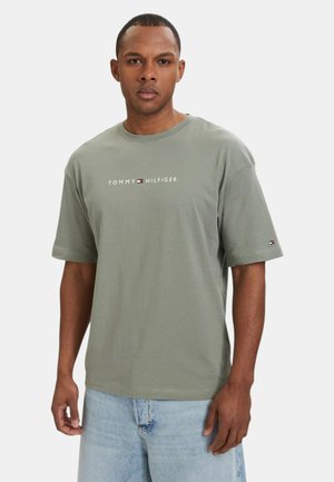 Man wearing a loose-fit olive green short-sleeve Tommy Hilfiger T-shirt with logo across chest and light blue denim jeans.