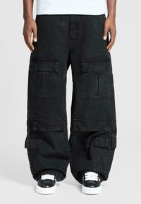 Persona che indossa pantaloni cargo neri oversize con molte tasche e sneakers bianco e nere su uno sfondo neutro.
