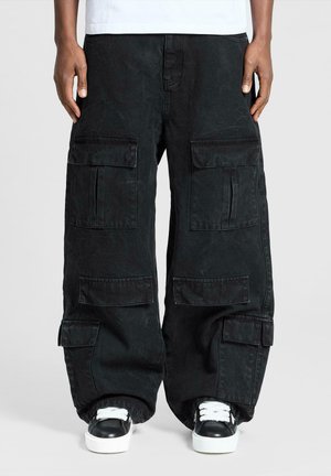 Personne portant un pantalon cargo noir surdimensionné avec plusieurs poches et des baskets noir et blanc sur un fond uni.