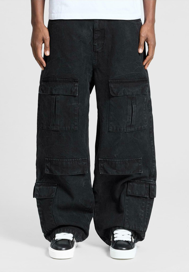 Persona che indossa pantaloni cargo neri oversize con molte tasche e sneakers bianco e nere su uno sfondo neutro.