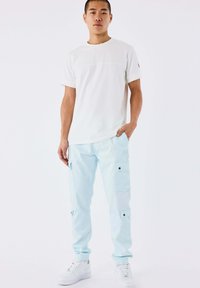 Giovane uomo in piedi, indossa una maglietta bianca, pantaloni cargo blu chiaro e sneakers bianche, con una mano in tasca, su sfondo bianco.