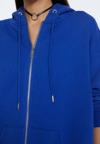 Felpa con zip blu realizzata in morbido tessuto, con cappuccio, accessori in argento, lacci e una tasca frontale. Accentato da un choker nero.