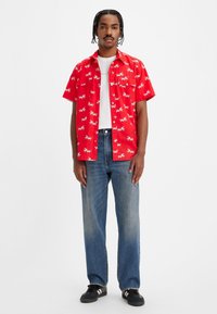 Hombre de pie que lleva una camisa corta de manga roja con estampado de hormigas blancas, camiseta blanca, jeans azules y zapatillas negras con rayas blancas.