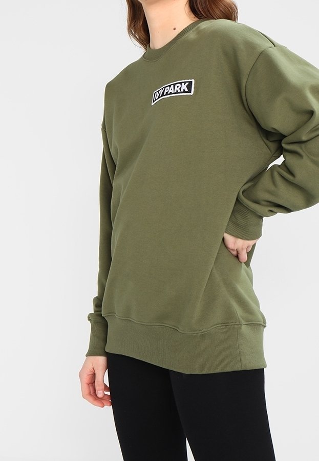 Person iført en olivengrøn langærmet sweatshirt med "IVY PARK" logo på brystet og sorte bukser, stående med den ene hånd på hoften.