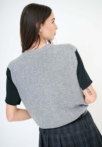 Maglione grigio senza maniche sopra una camicia nera. Il maglione ha una texture liscia, con scollatura arrotondata e vestibilità comoda. Gonna a quadri nera visibile.