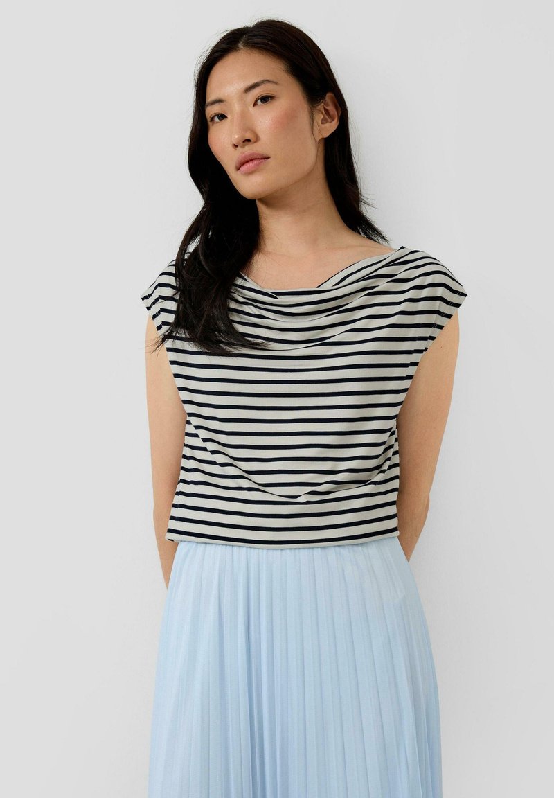 Vrouw met lang donker haar, gekleed in een zwart-wit gestreepte mouwloze top en een lichtblauwe geplooide rok, staand tegen een effen achtergrond.