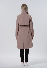 Lichtbruin waterdichte trenchcoat met een hoge kraag, een ceintuur bij de taille en knoopmanchetten; gedragen over zwarte broek en witte sneakers.