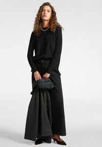Schwarzes Langarmoberteil, kombiniert mit schwarzen weit geschnittenen Hosen. Das Model hält eine puffy schwarze Jacke, die eine glatte Textur und einen glänzenden Finish aufweist.