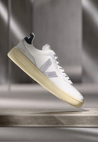 Sneaker bianca con tomaia in pelle liscia, accenti in gomma grigia, suola imbottita e un tab in pelle nera sul tallone. Presenta un logo e cuciture sottili.
