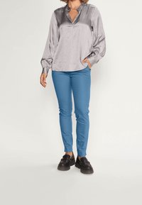 Blouse argentée soyeuse avec col en V et manches bouffantes, associée à un pantalon turquoise ajusté et des mocassins noirs épais.