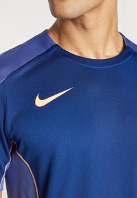 Camiseta deportiva azul de Nike con un tejido texturizado, acentos morados en los hombros y un logo de color naranja claro en el pecho.