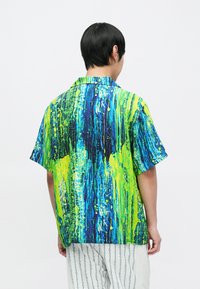 Koszula z krótkim rękawem w żywym niebieskim i neonowej zieleni z abstrakcyjnymi wzorami przypominającymi malowanie farbą. Charakteryzuje się luźnym krojem i klasycznym kołnierzykiem.