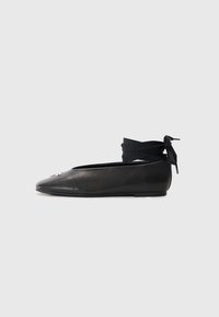Svart lærballerina med spiss tå, med et vevet stoffbånd rundt ankelen og en liten dekorativ detalj på tåen.
