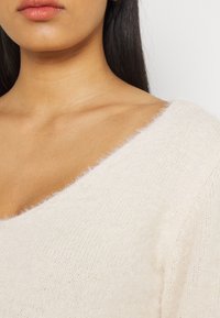 Cremfarbener, leicht flauschiger Pullover mit weitem V-Ausschnitt und strukturiertem Strick. Besitzt ein weiches, leichtes Material.