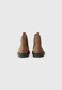 Bottines en suede marron avec des panneaux élastiques sur les côtés, des languettes de traction et des semelles en caoutchouc noir. Présentant une texture lisse et un design minimaliste.