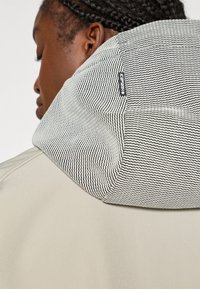 Μπεζ μπουφάν με υφή, ριγέ μαύρο και άσπρο καπέλο και μια μικρή ετικέτα λογότυπου στη ραφή του καπέλου. Το ύφασμα φαίνεται λείο και δομημένο.