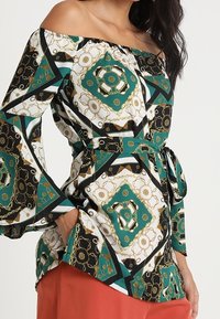 Donna che indossa un top off-shoulder con maniche a campana e stampa a catena geometrica nei colori verde, nero, bianco e oro, abbinato a pantaloni color ruggine.