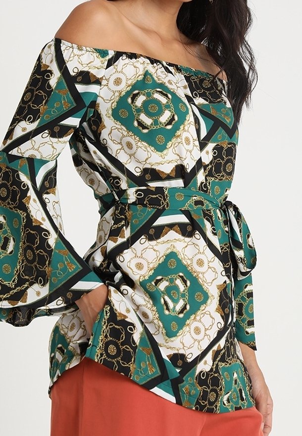 Donna che indossa un top off-shoulder con maniche a campana e stampa a catena geometrica nei colori verde, nero, bianco e oro, abbinato a pantaloni color ruggine.