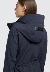 Giacca impermeabile blu navy con colletto alto, cappuccio regolabile con cordino e patch logo sul retro. Presenta un tessuto liscio e una vita elasticizzata. 