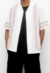 Kortærmet hvid skjorte med rød og sort broderet mønster langs skuldrene og lukningen; letvægtsstof, knaplukning foran.