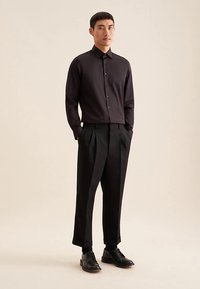 Camicia nera a maniche lunghe con colletto e bottoni sul davanti, abbinata a pantaloni neri a vita alta e scarpe nere su sfondo neutro.