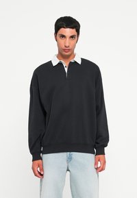 Svart hettegenser med hvit krage, to knapper foran og ribbestrikkede mansjetter, kombinert med lyseblå jeans. Uformell design, myk tekstur.