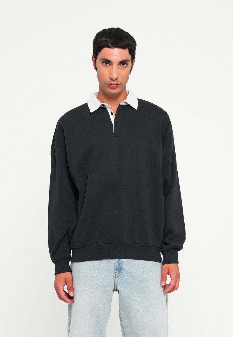 Svart hettegenser med hvit krage, to knapper foran og ribbestrikkede mansjetter, kombinert med lyseblå jeans. Uformell design, myk tekstur.