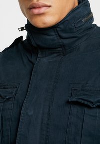 Marinblå jacka med hög krage, med texturerad tyg, framside dragkedja och två bröstfickor med klaffstängning.
