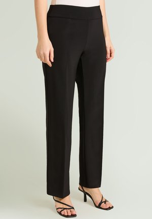 Kobieta w czarnych, prostych spodniach z wysokim stanem, w połączeniu z czarnymi sandałami na obcasie z paskami, stojąca na jasnym, jednolitym tle.