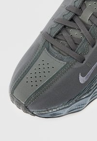 Close-up do bico de um sapatênis Nike cinza-escuro com couro perfurado, parte superior em tecido, atacadores cinzentos e logótipo swoosh branco na lateral.