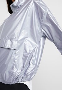 Helle lila Windjacke mit hohem Kragen, elastischen Bündchen und einer Fronttasche. Hergestellt aus einem glänzenden, leichten Material mit geriffelter Textur.