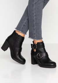 Schwarze Knöchelstiefel mit einem klobigen Absatz, ausgestattet mit einem seitlichen Reißverschluss, zwei breiten Riemen und runden Hardware-Akzenten an den Seiten.