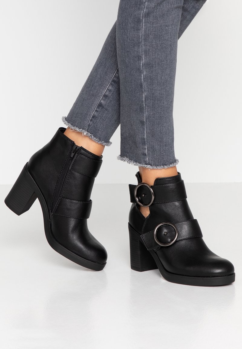 Schwarze Knöchelstiefel mit einem klobigen Absatz, ausgestattet mit einem seitlichen Reißverschluss, zwei breiten Riemen und runden Hardware-Akzenten an den Seiten.