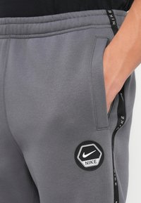 Pelēkie sporta bikses ar mīkstu tekstūru, ar melnu Nike logo plāksteri, sānu kabatām un melnām zīmola strīpām pa malām.