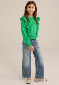 Pulover verde tricotat cu mâneci franjurate, linii verticale texturate; asortat cu blugi largi de culoare albastru deschis și pantofi bej.