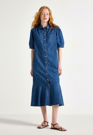 Kvinde med langt rødt hår iført en blå denim midi-kjole med knapper foran, puffede korte ærmer og brune sandaler, står mod en ensfarvet baggrund.