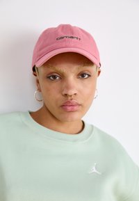 Gorra rosa con el logotipo bordado de "Carhartt", combinada con una sudadera verde menta que presenta un logotipo blanco. Tejido suave y corte relajado.