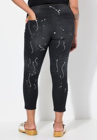 Pantalones vaqueros skinny de mezclilla negra con patrón de salpicaduras blancas, dos bolsillos traseros, de longitud hasta el tobillo y ligero desgaste en la textura.