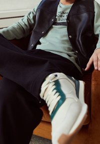 Vit sneaker med teal accenter och texturerade snören, bärs med mörka sweatsbyxor och en ljusgrön tröja, sitter på en orange stol.