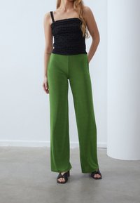 Kvinna klädd i svart rynkad tanktop och gröna vida byxor, stående inomhus på grått golv med händerna bakom ryggen.