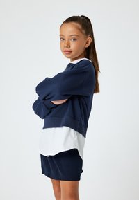 Granatowy sweatshirt z obszernymi rękawami, nałożony na białą koszulę z kołnierzykiem. Połączony z granatowymi szortami, gładka tekstura, casualowy design.