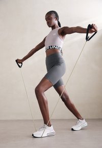 Donna atletica che utilizza bande di resistenza, indossando un crop top rosa chiaro e pantaloni grigi, con scarpe da ginnastica bianche, in un ambiente interno.