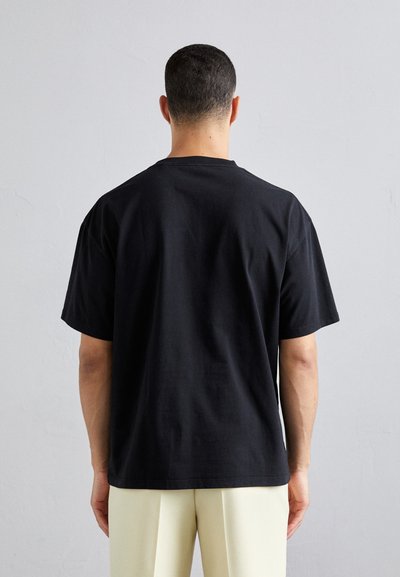 Sort kortærmet t-shirt lavet af bomuld, med en afslappet pasform og rund udskæring, båret med lyse bukser.