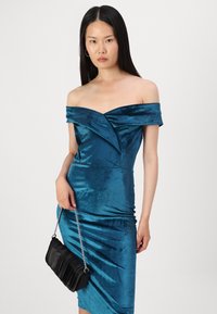 Tirkizna halter haljina od baršuna s pripijenom siluetom, koja ima drapirane detalje na bustu i elegantnu, teksturiranu površinu. Crna lančana torba.