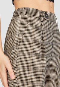 Pantalon taille haute à carreaux avec un motif à carreaux beige et noir, tissu léger, texture lisse, et fermeture avant par bouton.