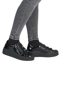 Ben i grå skinny jeans iført skinnende sorte plateau-sneakers med brede båndsnørre og sidelommer med lynlås, mod en hvid baggrund.