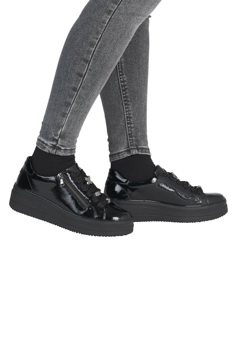 Ben i grå skinny jeans iført skinnende sorte plateau-sneakers med brede båndsnørre og sidelommer med lynlås, mod en hvid baggrund.