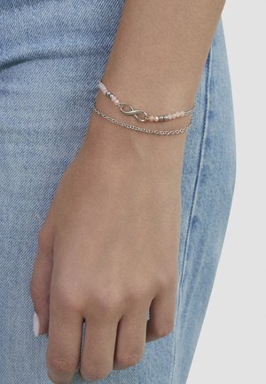 Hand resting by side wearing two bracelets: one with silver infinity symbol and pink beads, the other a simple silver chain, against blue denim.