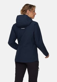 Una donna che indossa una giacca con cappuccio Mammut di colore blu navy e pantaloni neri, in piedi e rivolta verso destra, su uno sfondo neutro.